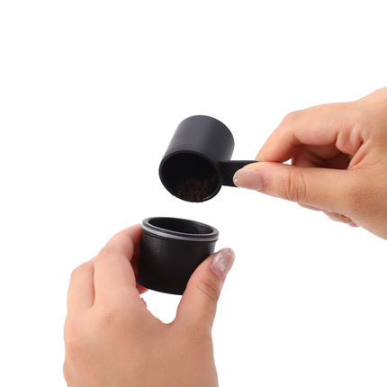 Black coffee scoop being used to transfer coffee grounds into a container held by hands on a white background