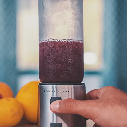 Hand holding a blender with a purple smoothie, surrounded by fruits on a blurred background