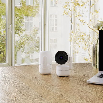 White robot vacuum cleaner on a wooden floor with a laptop and decorative plant in the background.