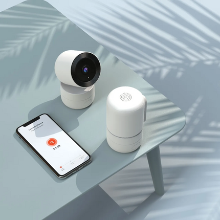 Smart home devices including a camera, speaker, and smartphone with app interface on a glass table.